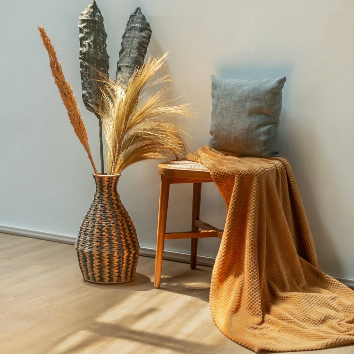 Boho vaas DAYANA van waterhyacint met gedroogde bloemen naast houten stoel met oranje deken en blauw kussen in zonlicht.