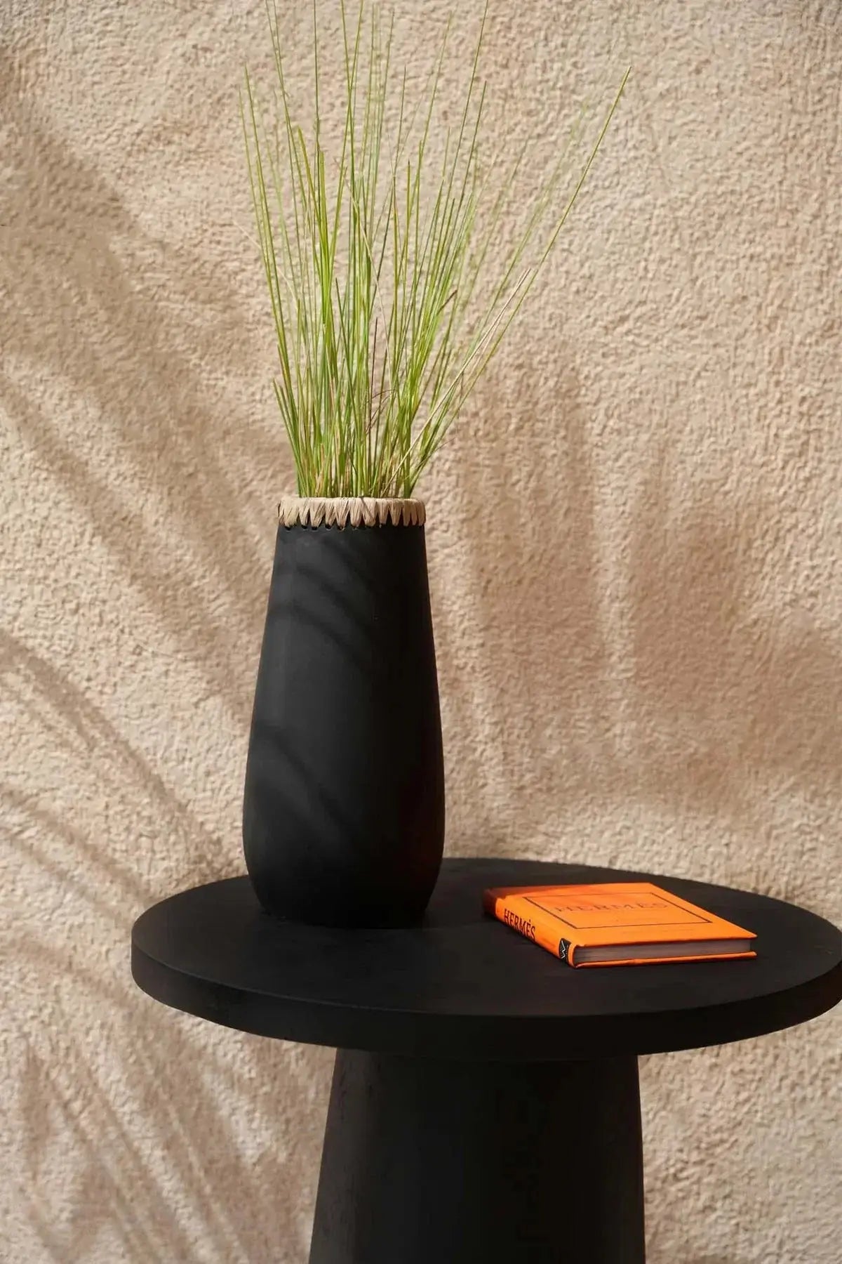 Zwarte Boho vaas The Sneaky L op ronde tafel, elegant handgemaakt design met groene plantenstengels, perfecte aanvulling voor modern interieur.