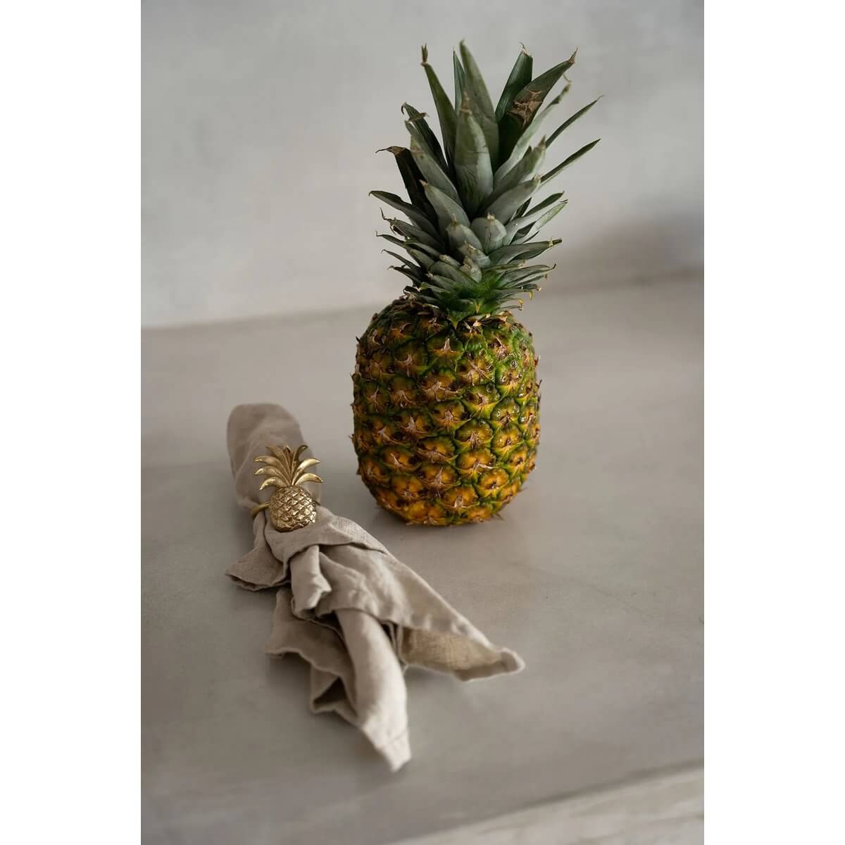 Bohemian-style table setting with gold pineapple napkin ring and actual pineapple on neutral surface for luxurious dining ambience.