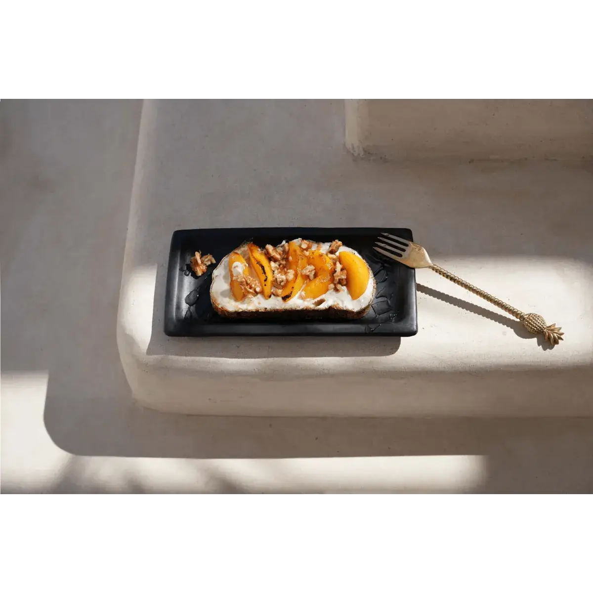 Golden bohemian pineapple fork beside a dessert on a black plate, casting elegant shadows.