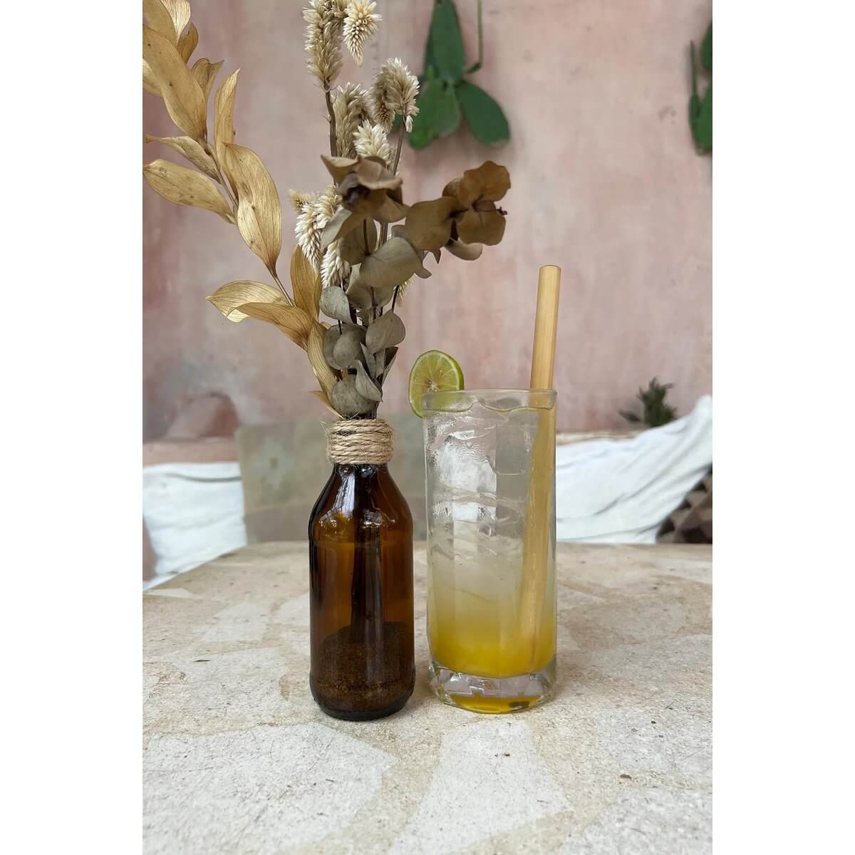 Glass with iced cocktail and bamboo straw beside a rustic brown vase with dried flowers, set against a Bohemian decor backdrop.
