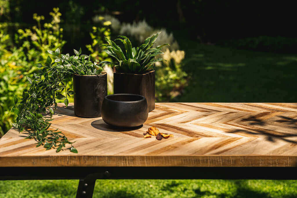 Houten tafel met zwarte teakhouten potten en groene planten in een zonnige tuinomgeving.