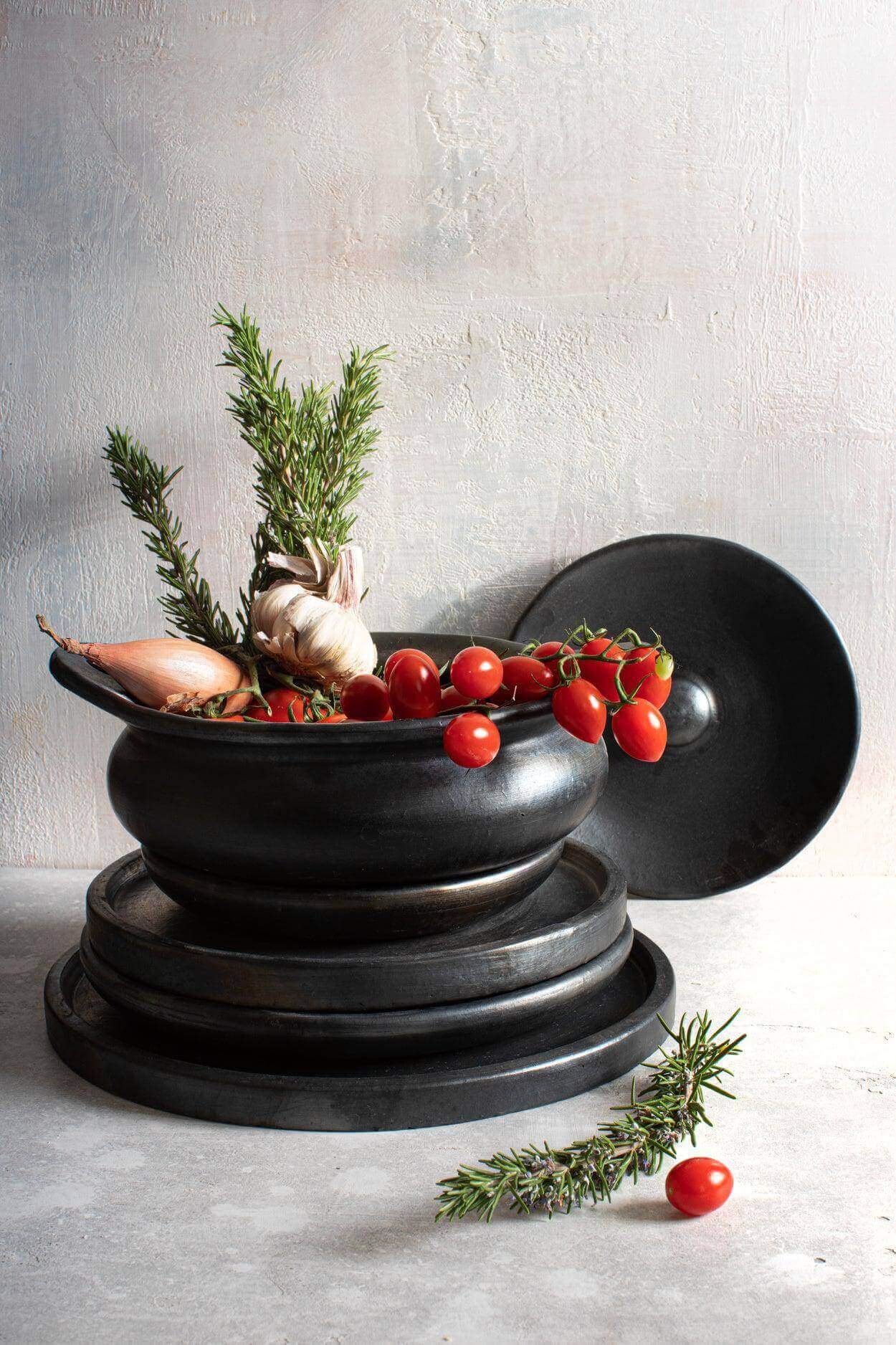 Elegant zwarte serveerpot van gebrande terracotta met deksel, gevuld met verse kruiden en tomaten, perfect voor een stijlvolle tafel.