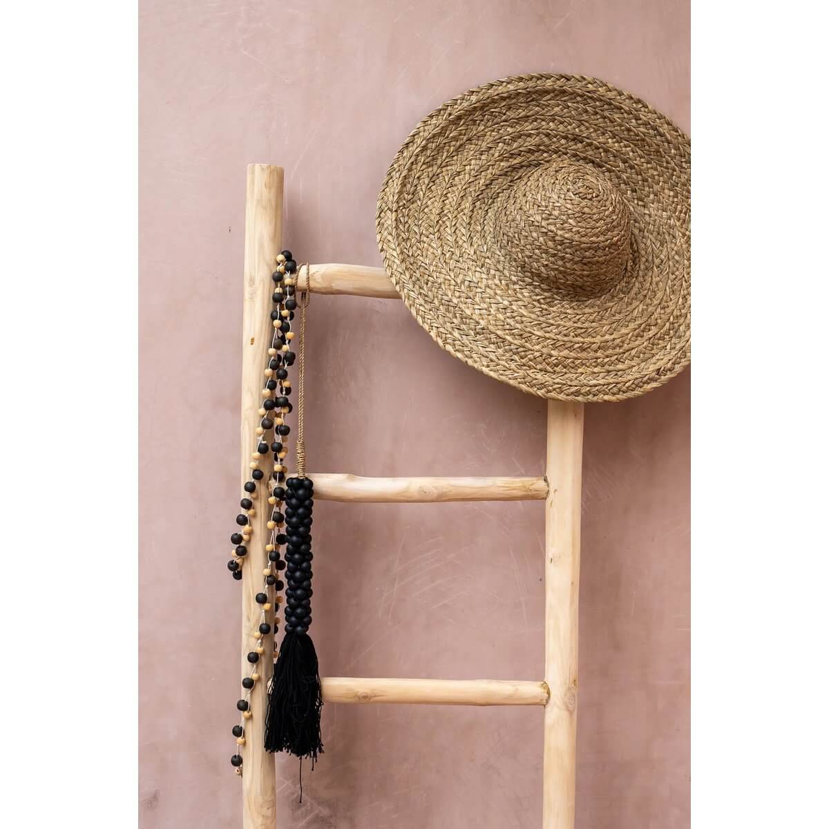 Bohemian De Canggu ketting in natuurlijk zwart hangt aan een houten ladder, naast een strohoed tegen een roze muur.