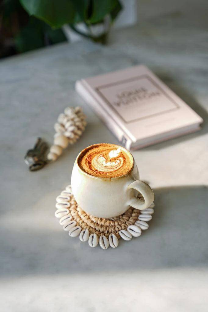 Witte koffiemok met latte art op een bohemian onderzetter, naast een boek op een zonnige tafel.