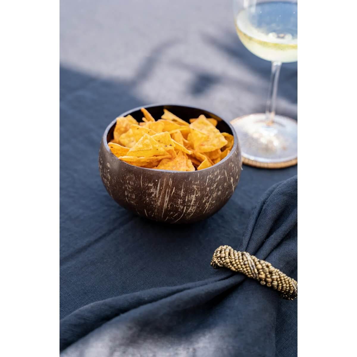 Bohemian style coconut bowl filled with chips on a black tablecloth, accompanied by a glass of white wine.