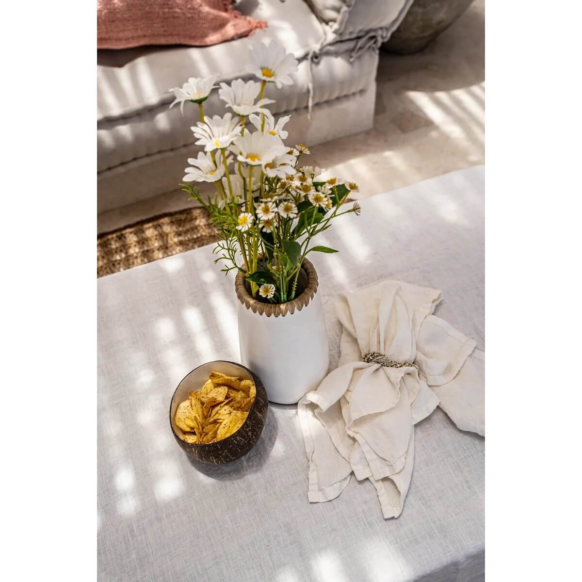 Bohemian decor with white daisies in a vase, coconut bowl of chips, and elegant napkin on a cozy table setting.