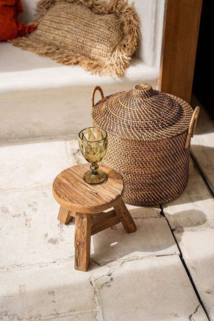 Koloniale rotan mand met deksel en handvaten, naast rustiek houten krukje en glas, op witte vloer. Perfecte opslagoplossing.
