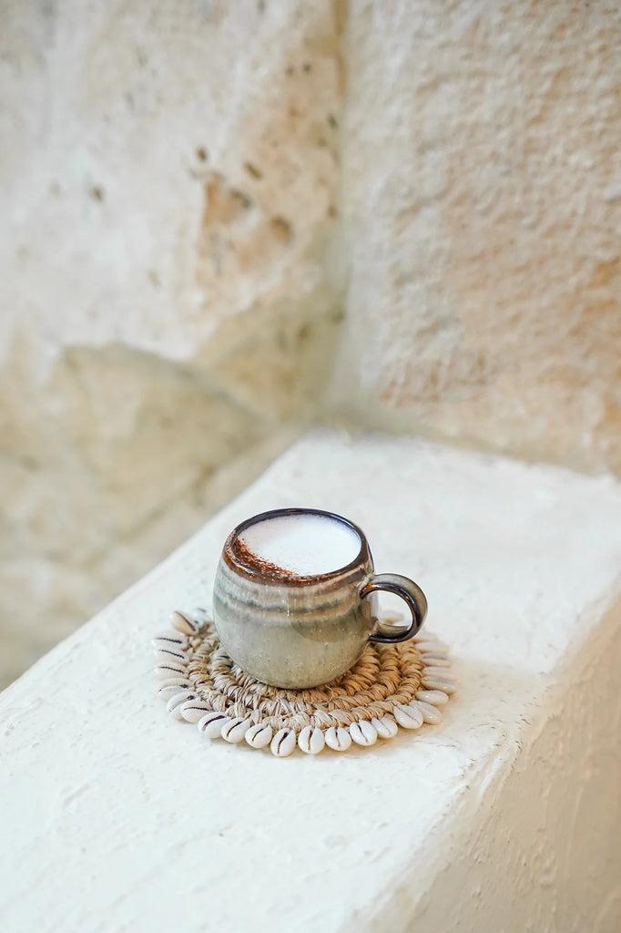 Handgemaakt aardewerk espresso kopje op decoratieve onderzetter tegen een rustieke muur, perfect voor brunches of diners.