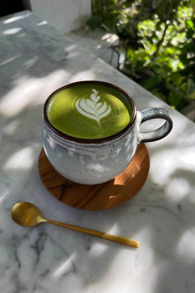 Een matcha latte in een mooi aardewerken kopje met latte art, op een marmeren tafel met een gouden lepel ernaast.