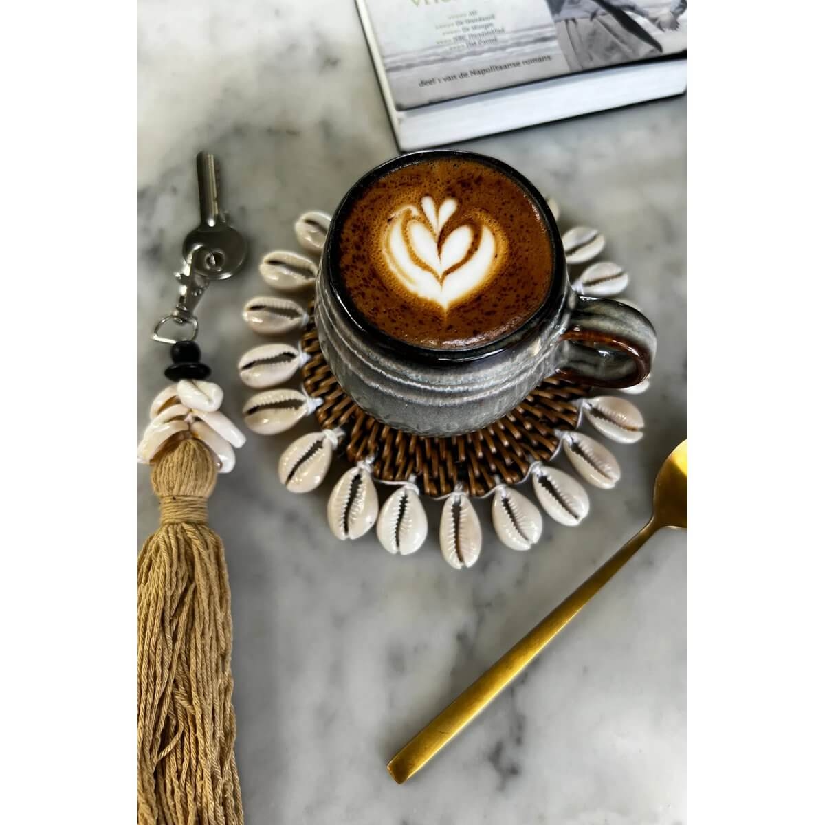 Alt Tag: Bohemian De Cowrie Tassel Keychain in Mocca beside a coffee on shell coaster, golden spoon nearby on marble surface.