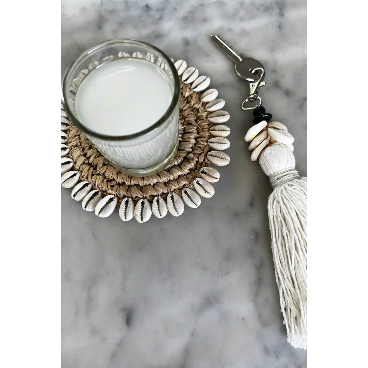 Bohemian De Cowrie Tassel Keychain in white with shells and cotton tassel next to a glass of milk on a decorative mat.