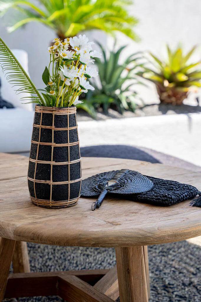 Elegante zwarte terracotta vaas met rotan, gevuld met bloemen, op houten tafel in een zonnige buitenruimte.