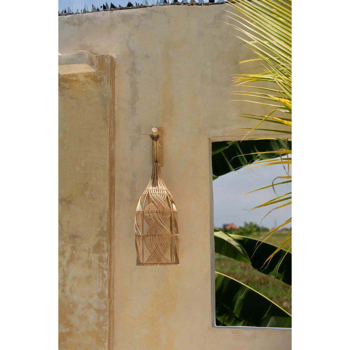 Alt Tag: Rotan De Garaffa-hanger lamp in natureel, opgehangen tegen een beige muur buiten met een spiegel, geschikt voor diverse lampstijlen.
