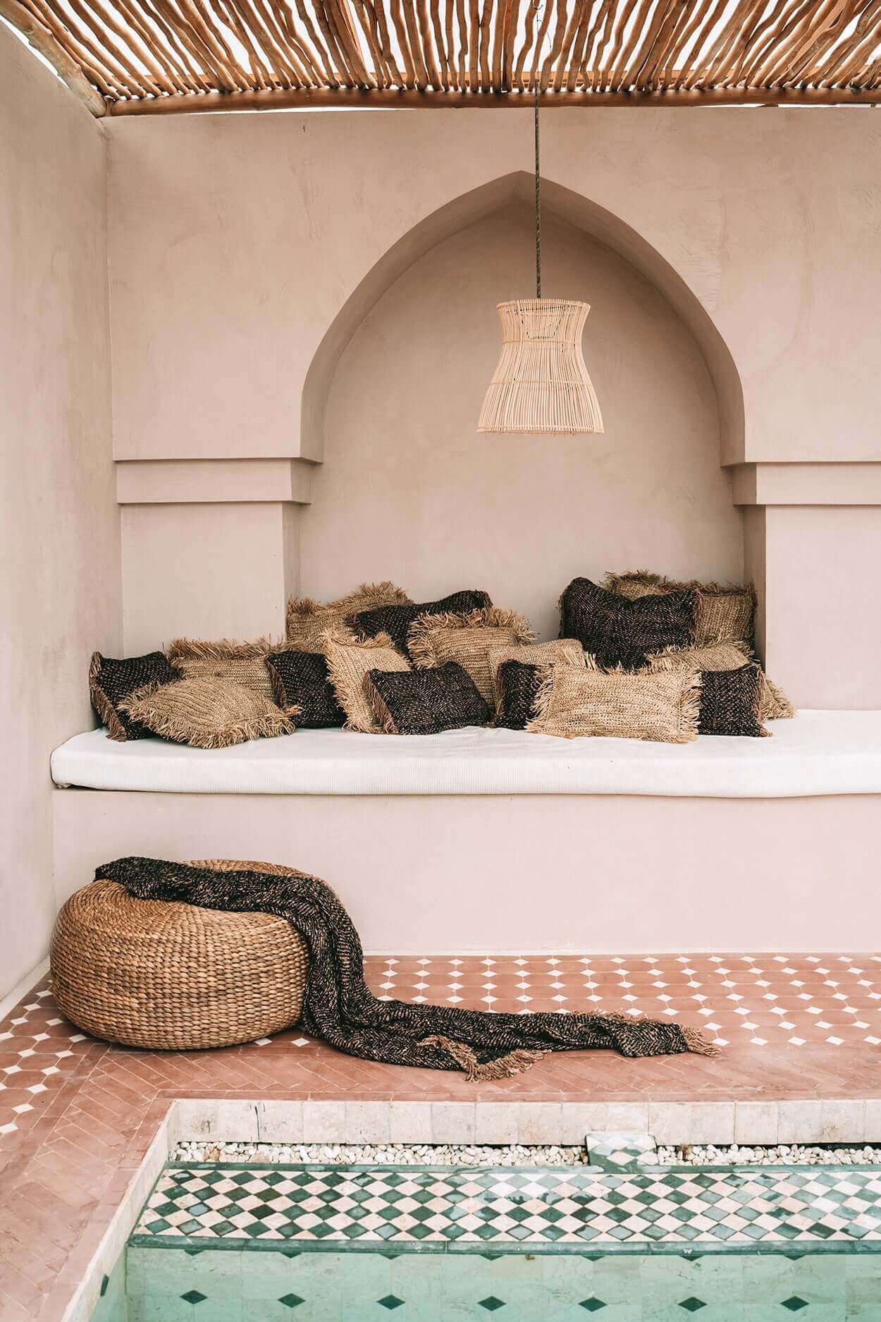 Stijlvolle boho zithoek met handgemaakte poef en kussens, naast een zwembad en onder een rieten lamp. Perfect voor tuin of terras.