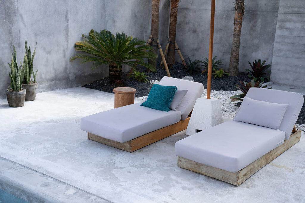 Twee comfortabele ligbedden met kussens naast een zwembad in een moderne buitenruimte met planten en een parasol.