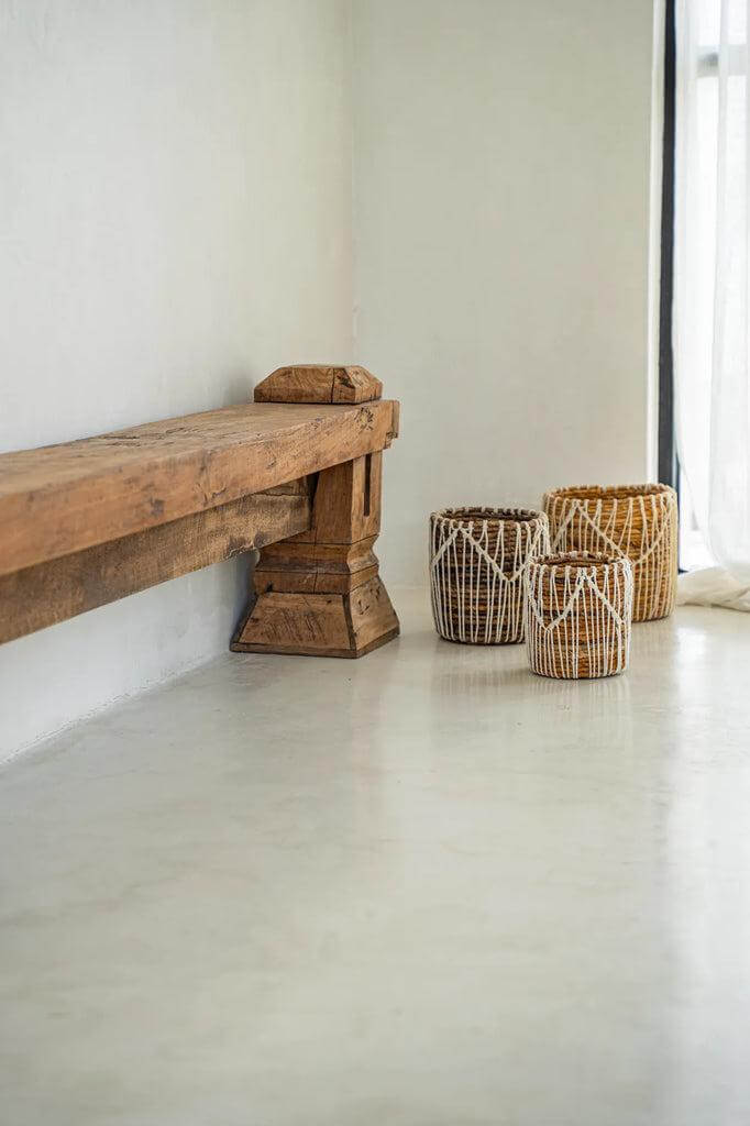Drie handgemaakte macramé manden van bananenbladeren in aardetinten naast een rustieke houten bank op een gladde vloer.