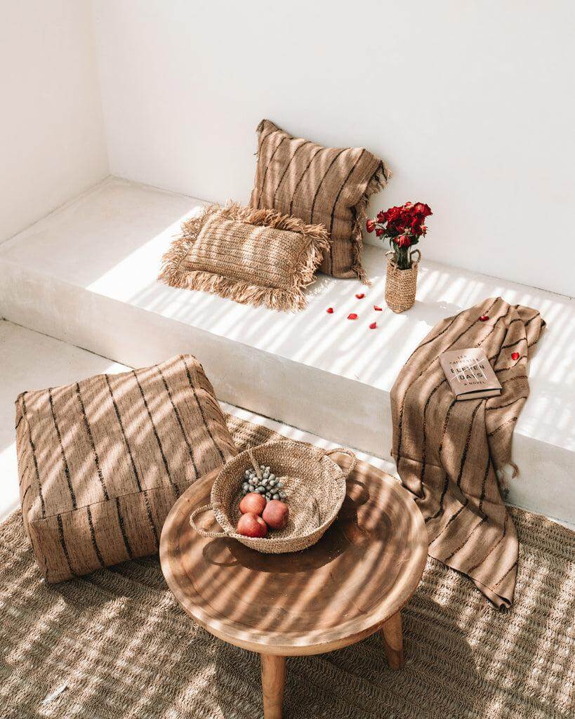 Boho hoek in de zon met beige en zwarte kussens, rotan poef en mand, ideaal voor een chique en ontspannen sfeer. Perfect voor binnen of buiten.