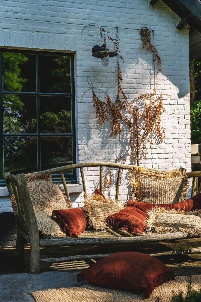 Gezellige tuinbank met De Oh My Gee kussenhoes in bordeaux fluweel, boho woondecoratie bij een wit bakstenen huis.