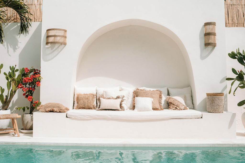 Stijlvolle buitenruimte met witte bank en kussens bij een turquoise zwembad, omgeven door tropische planten en elegante muurverlichting.