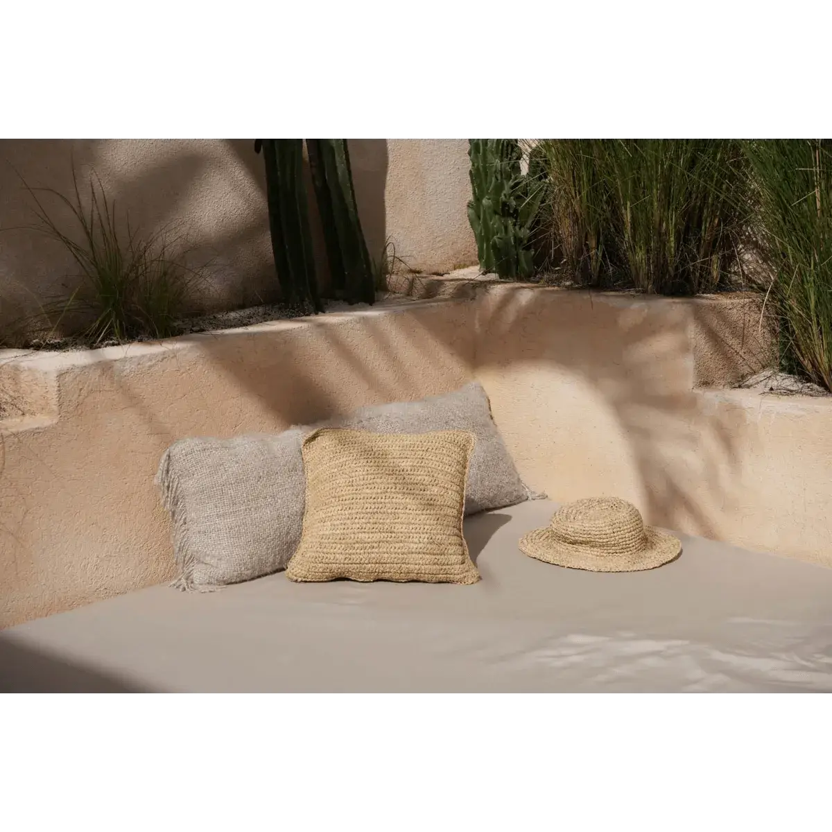 Bohemian De Pantai-hoed naast kussens op een zonnig terras, perfect voor stranddagen. Gemaakt van zeegras, stijlvol en lichtgewicht.