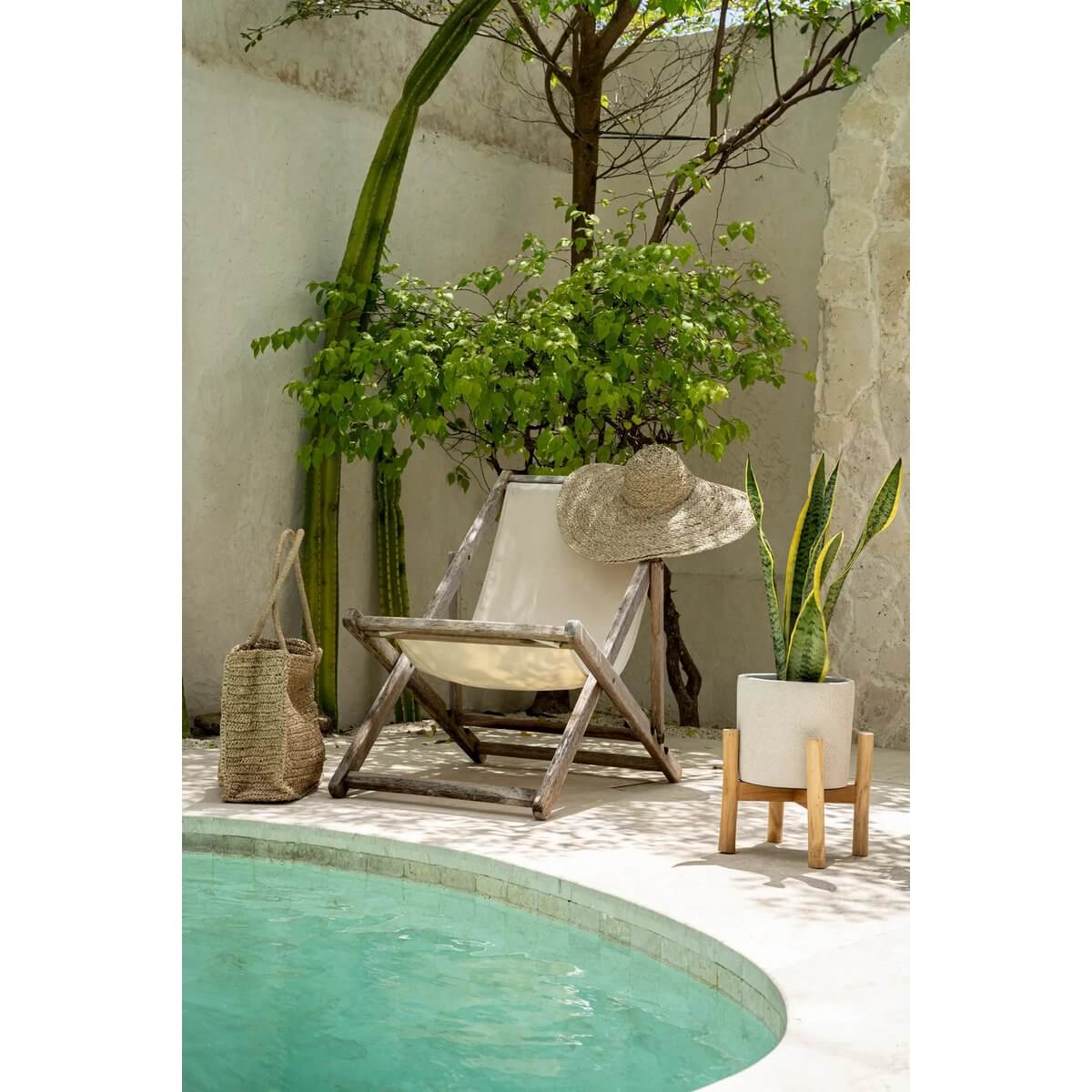 Cozy bohemian poolside scene with a rustic chair, De Playa hat, woven bag, and potted plant in a serene outdoor setting.