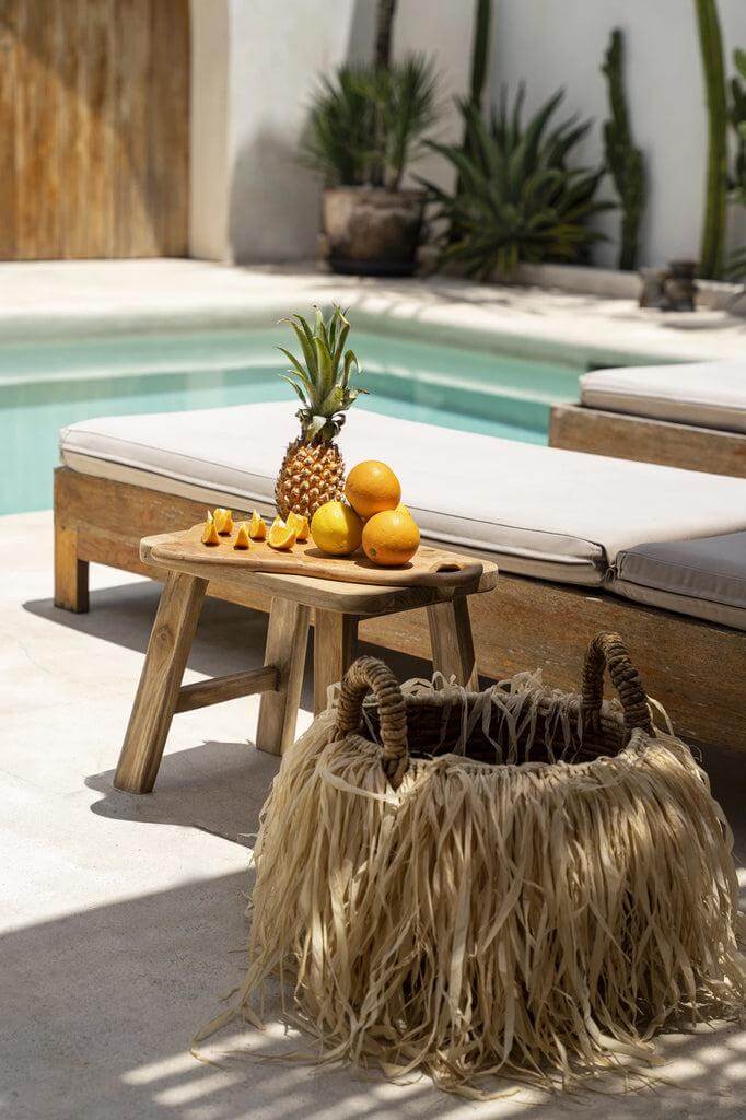 Houten bijzettafel met ananas en sinaasappelen bij een zwembad, naast een decoratieve mand met grasachtig materiaal.