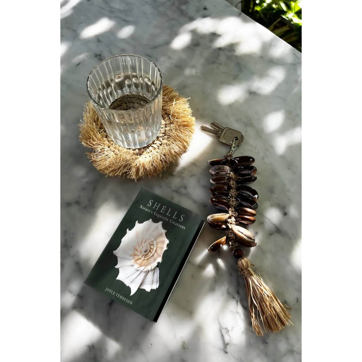 De Raffia Fishbone Keychain in natural black with bohemian charm lies on a marble surface beside a glass and a seashell book.