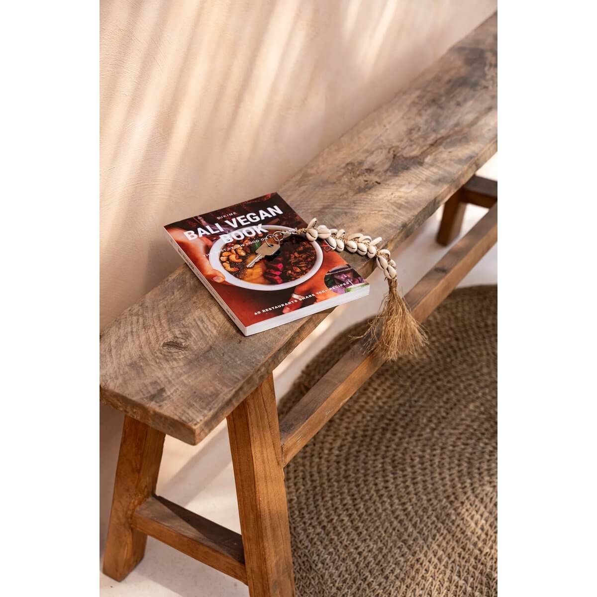 Bohemian De Raffia Fishbone Keychain in natural white on wooden bench beside Bali Vegan cookbook.