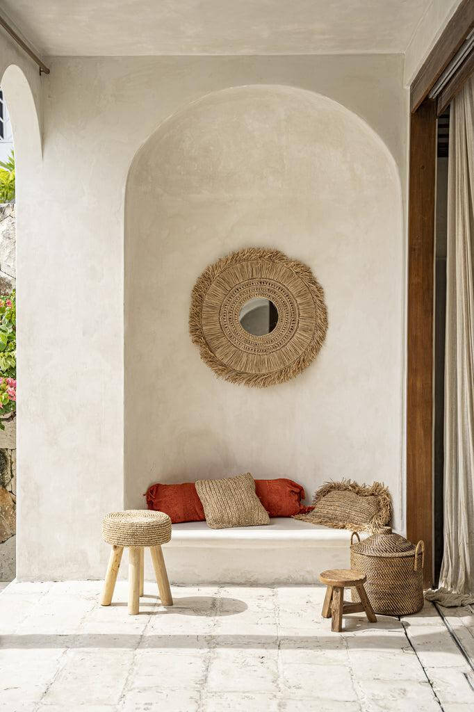 Rustieke zithoek met natuurlijke accenten, inclusief De Raffia Kruk, gevlochten spiegel en terracotta kussens. Warme, gezellige sfeer.