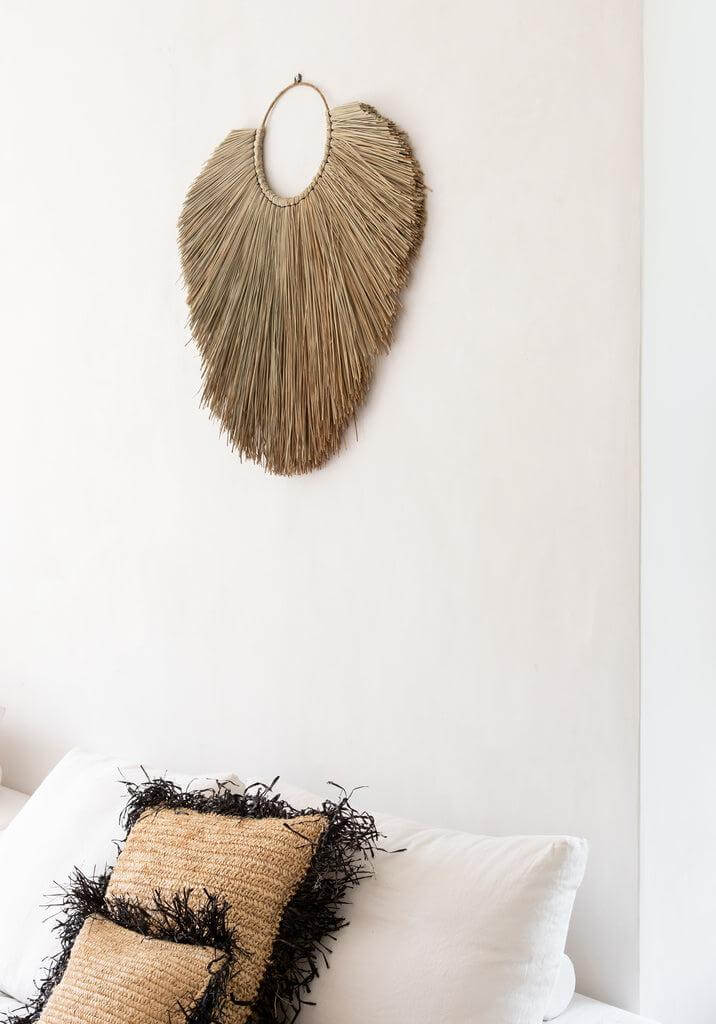 Raffia kussenhoes in natuurlijk zwart op witte bank met decoratieve wandhanger, perfect voor boho interieurstijl.
