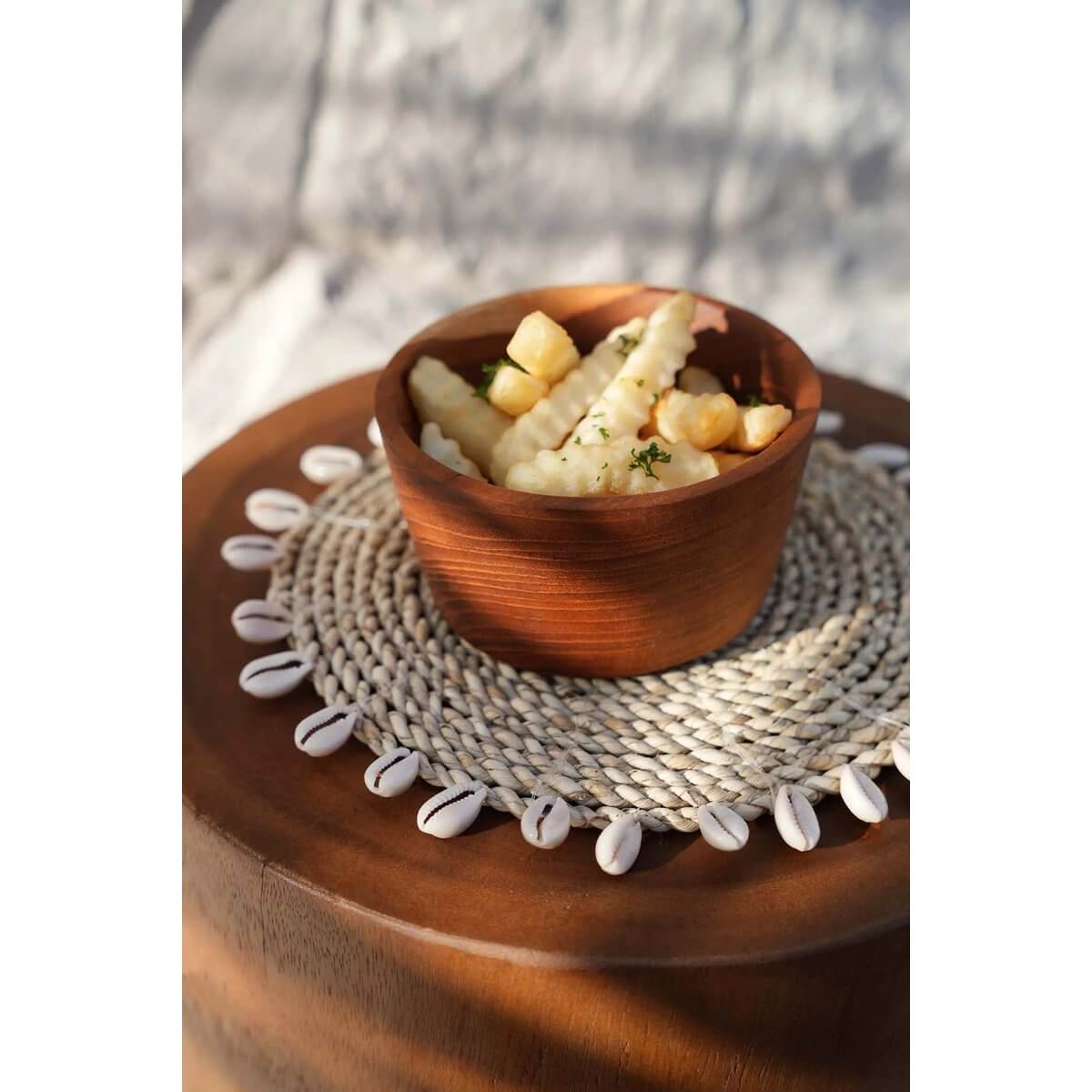 Bohemian raffia and shell pot holder with kauri detailing, showcasing a wooden bowl on a rustic table. Perfect island-style accent.