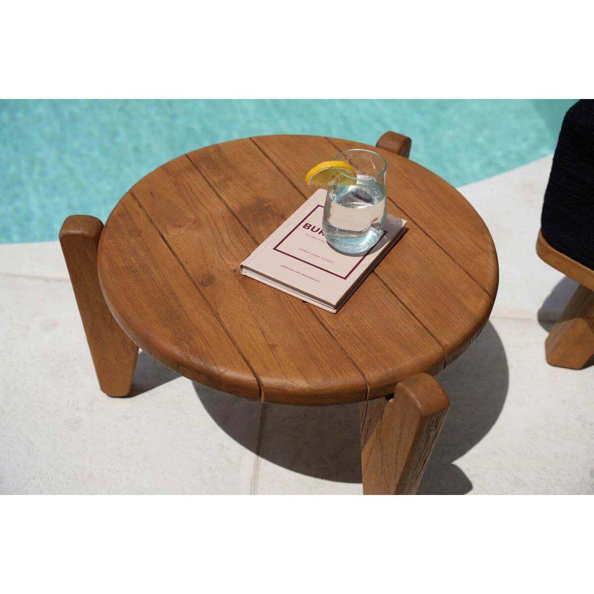 Ronde teakhouten Seseh-salontafel met drie poten naast zwembad, met boek en glas water erop.