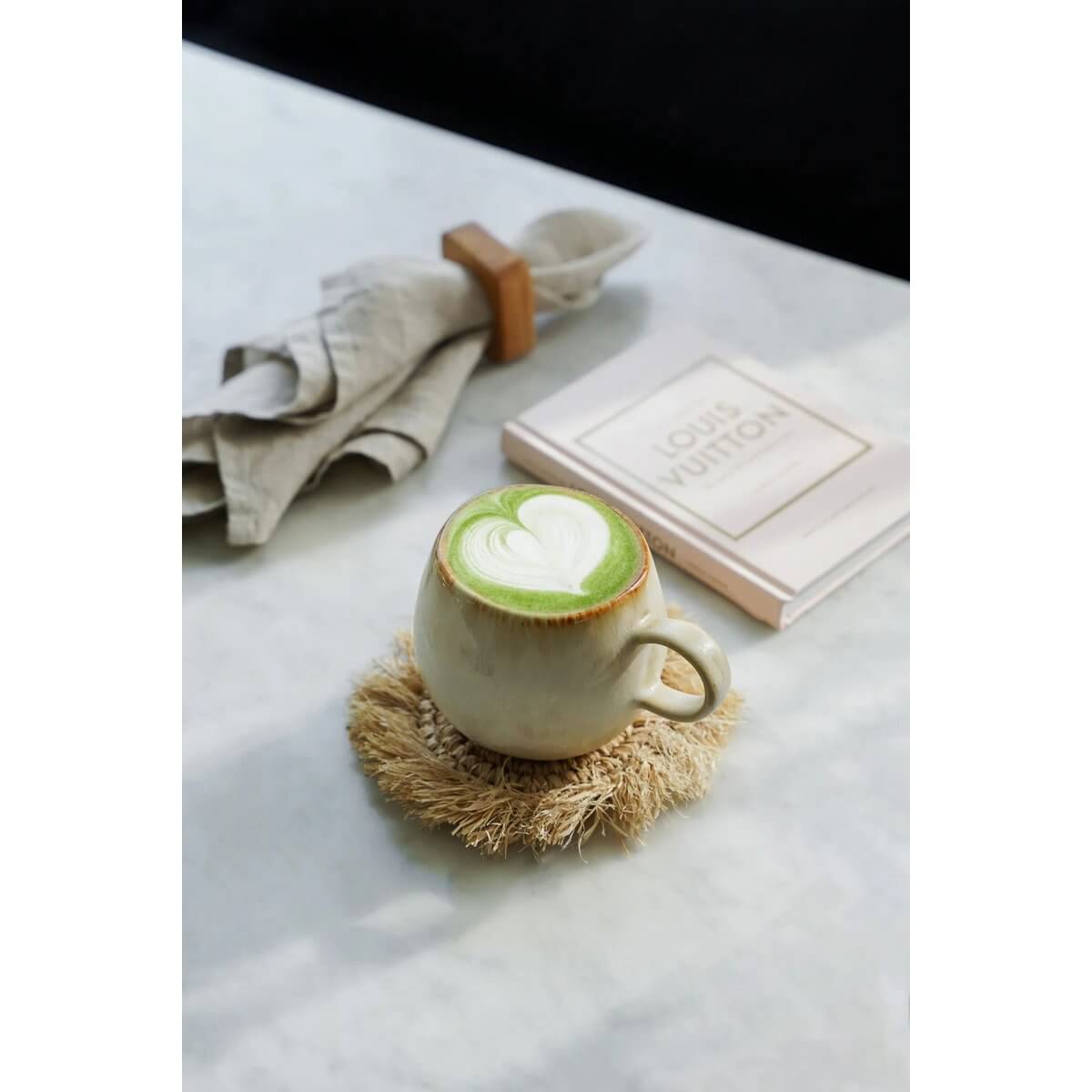Matcha latte with heart foam art in ceramic mug, resting on straw coaster, elegant beige napkin and book in background. Cozy bohemian vibe.