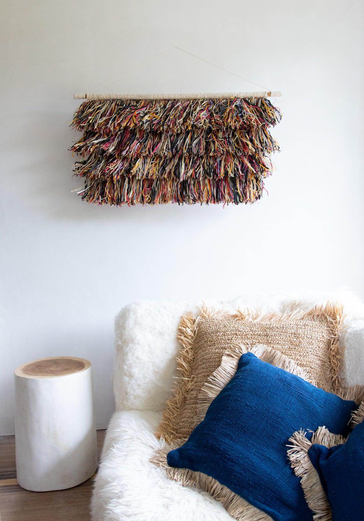 Witte houten kruk naast een rustieke bank met kleurrijke wanddecoratie en blauwe kussens in een gezellige woonkamer.