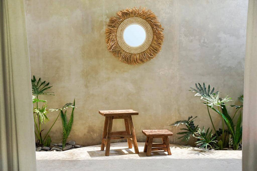 De Yoke krukken in twee maten, handgemaakt van gerecycled teakhout, tegen een rustieke muur met spiegel en groene planten, stijlvolle sfeer.