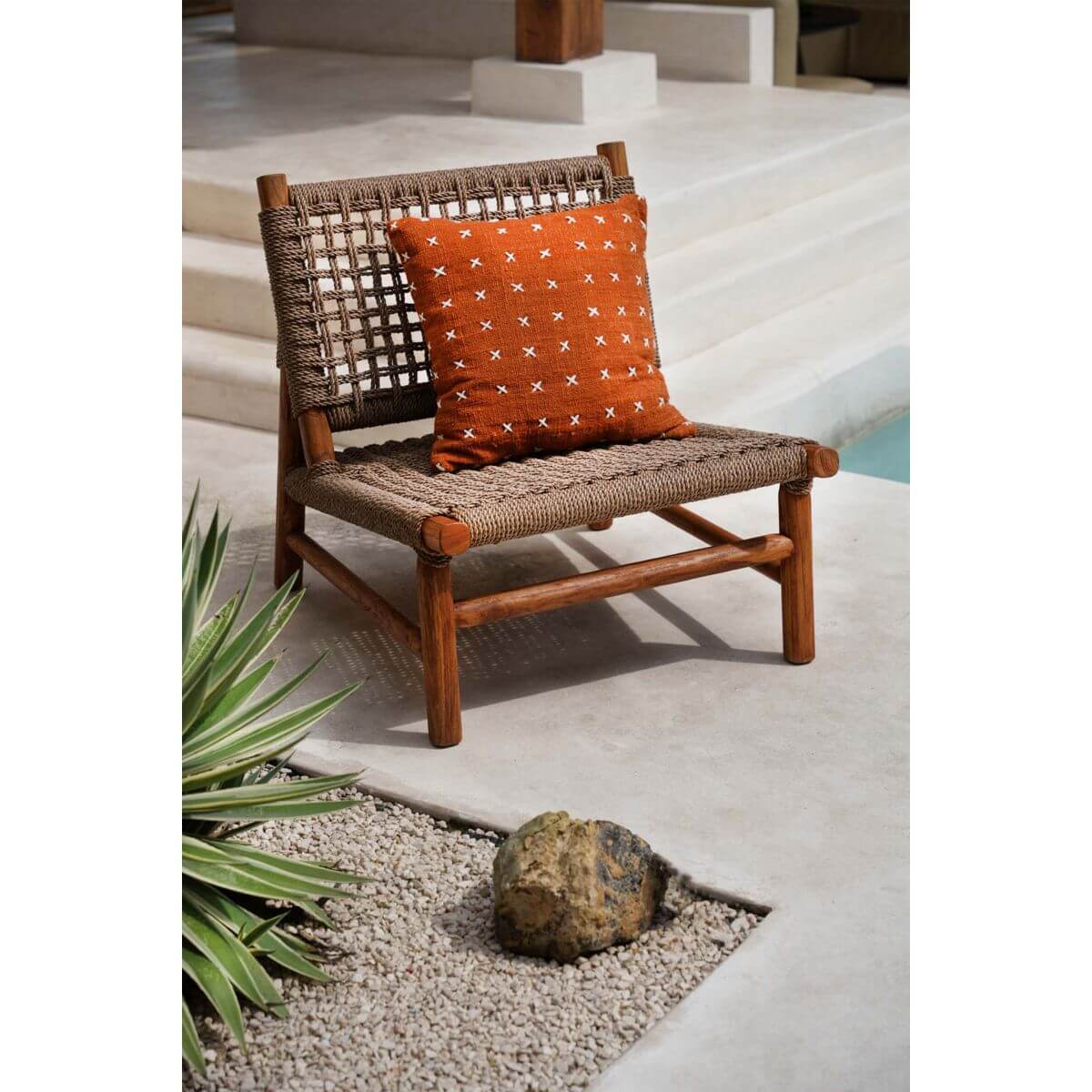 Rotan stoel met terracottakleurige Zillion Kiss kussenhoes, naast een zwembad met groene planten en natuurlijke stenen.