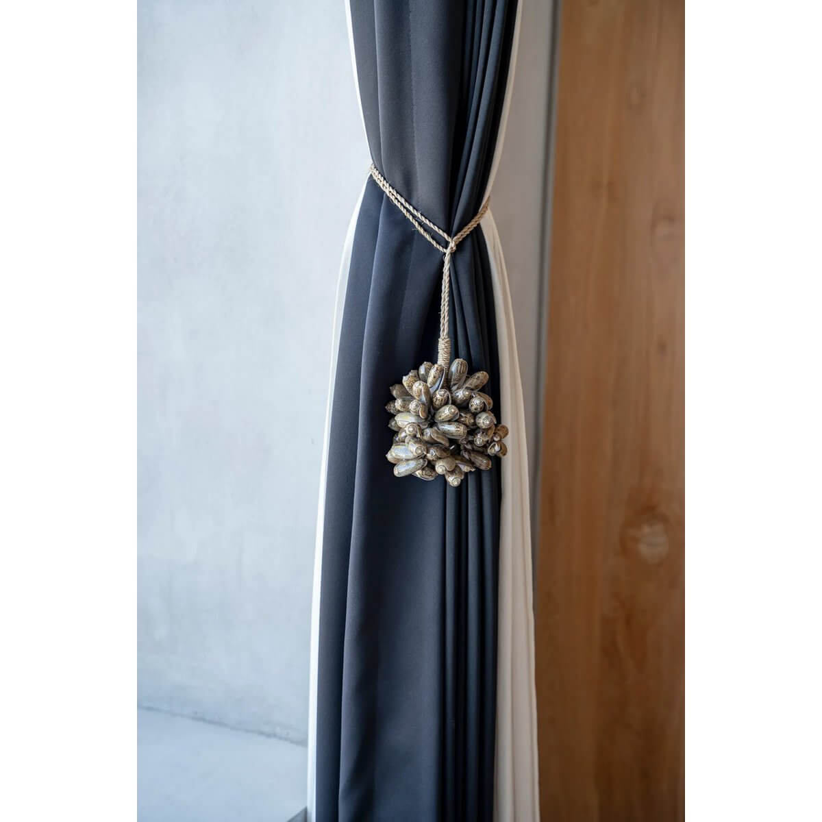 Handmade brown kauri shell tassel hanging on a curtain, adding a bohemian touch to interior decor. By Bazar Bizar Living.