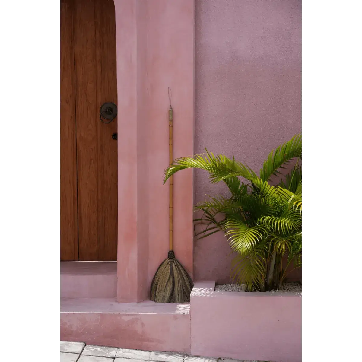 Bohemian-style Indonesian grass broom in black, gray, and gold, leaning against a pink wall beside green plants.