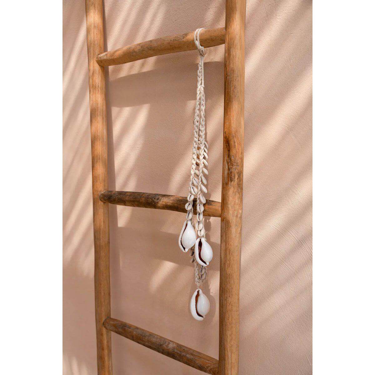 De grote witte kauri schelp ketting aan een houten ladder, voegt een vleugje kustflair en bohemien charme toe aan de woonruimte.