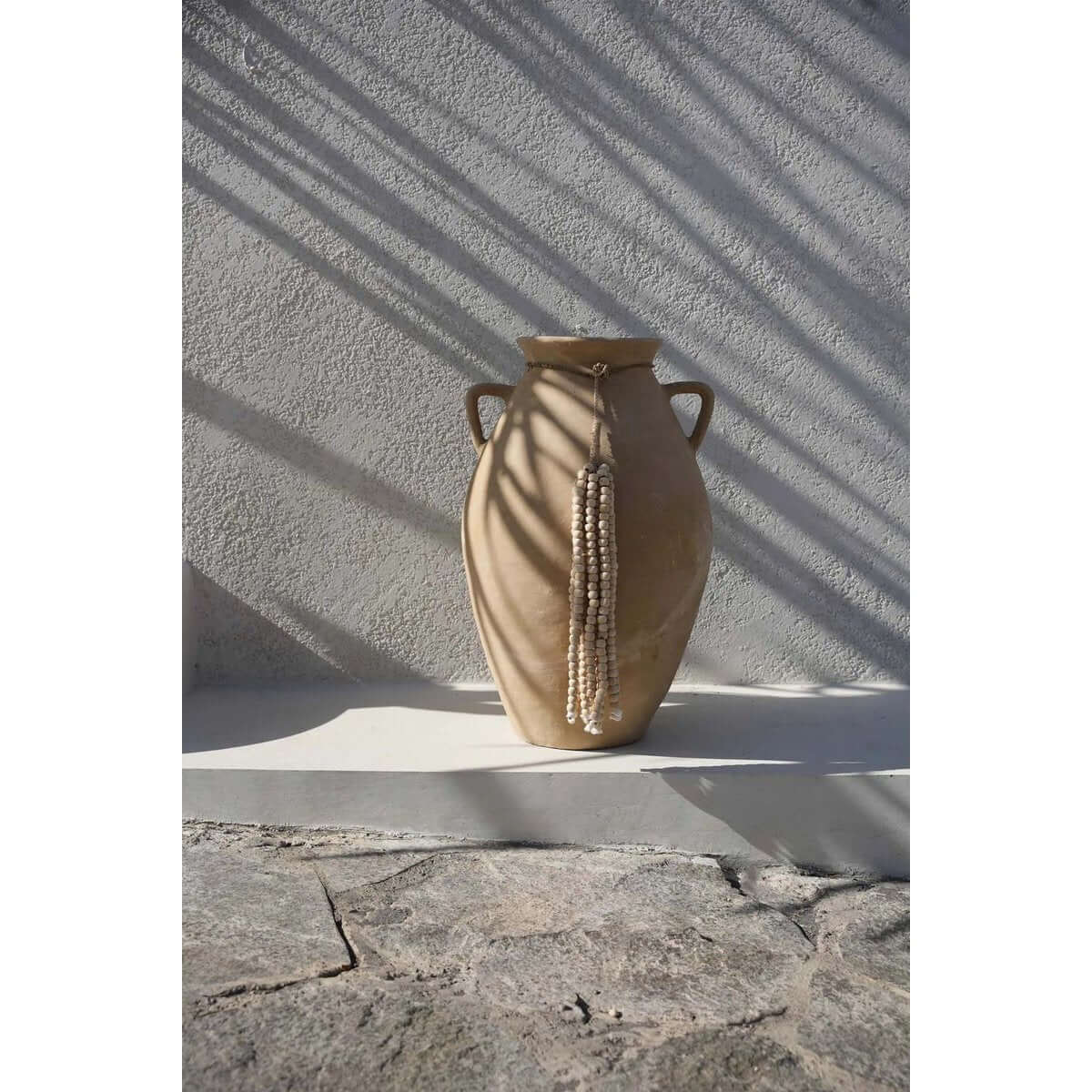 De houten kralen kwast - naturel bohemian decor hanging on traditional clay vase, casting shadows on rustic surface.