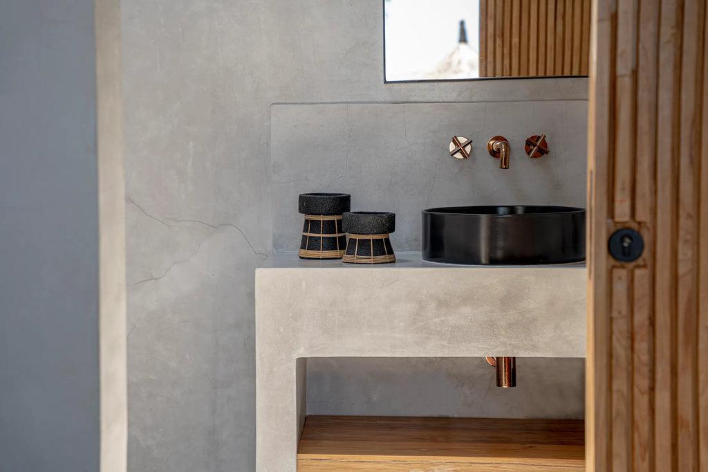 Twee zwarte terracotta kaarsenhouders met rotanrooster op een minimalistische badkamer wastafel.