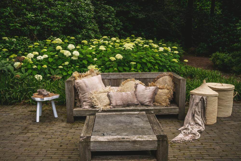 Houten bank met kussens en plaid in een gezellige tuinomgeving met weelderige groene struiken en bloemen op de achtergrond.