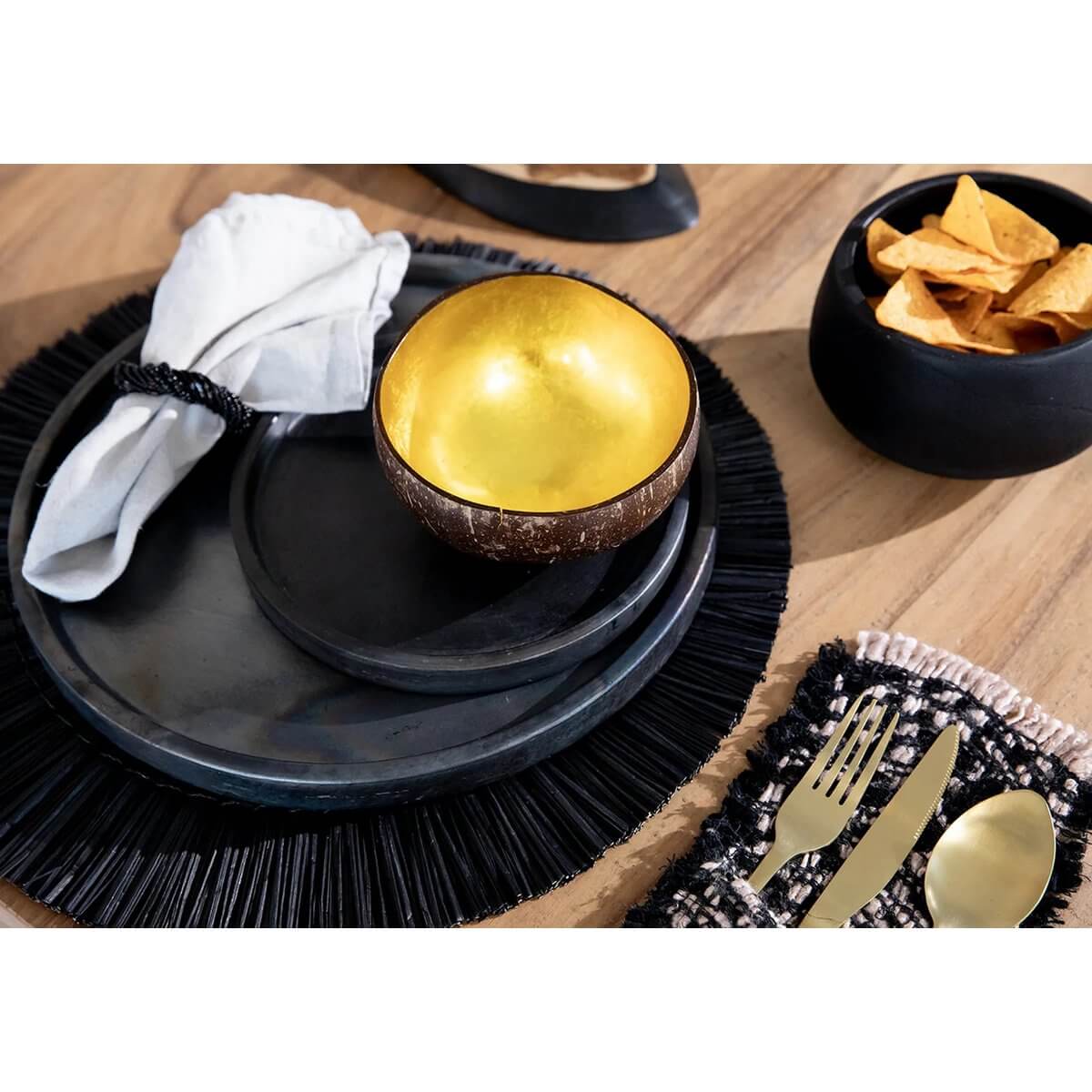 Bohemian table setting with black ceramic plates, gold coconut bowl, and napkin on wooden surface. Stylish and elegant dining ware.