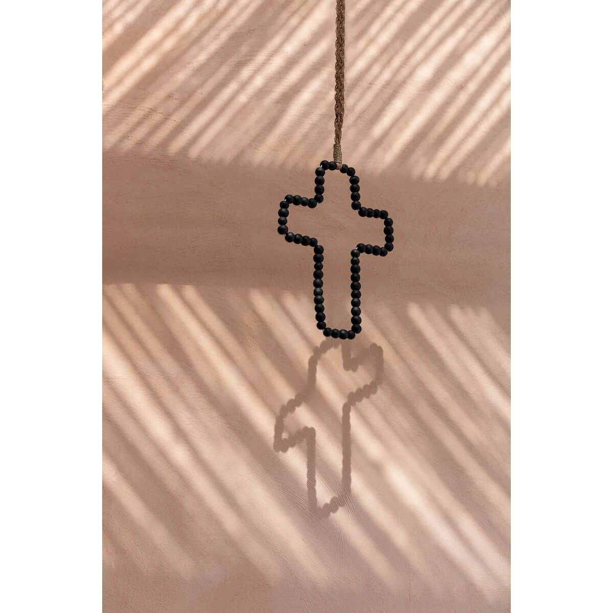 Bohemian wooden cross with beads in black and grey, casting a shadow on a textured wall. Handcrafted decor by Bazar Bizar Living.