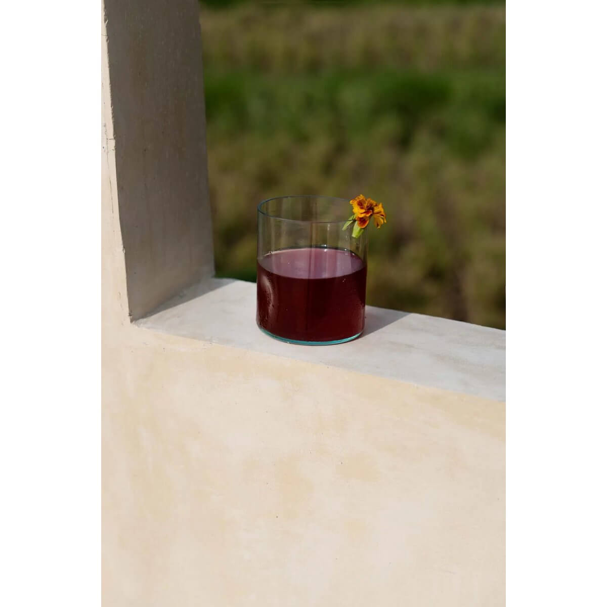 Bohemian style recycled glass on block, filled with rich dark liquid and flower garnish, surrounded by lush green field for eco-friendly vibe.