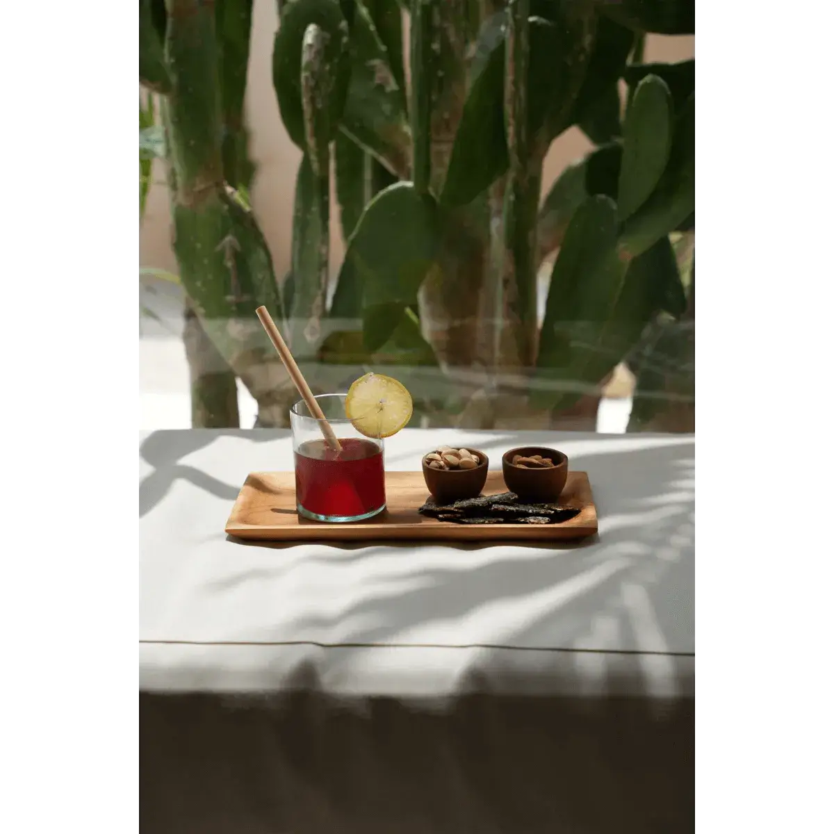 Bohemian recycled glass with red drink and lime slice, two treats on wooden tray, cactus background, eco-friendly styling.