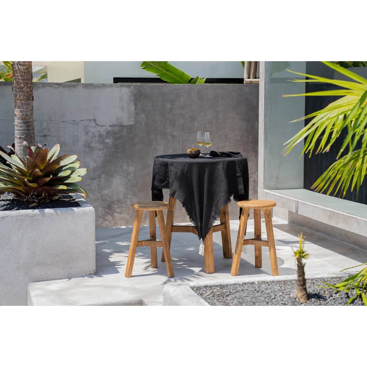 Buitentafel met zwart linnen tafelkleed en houten krukken, omringd door palmen.