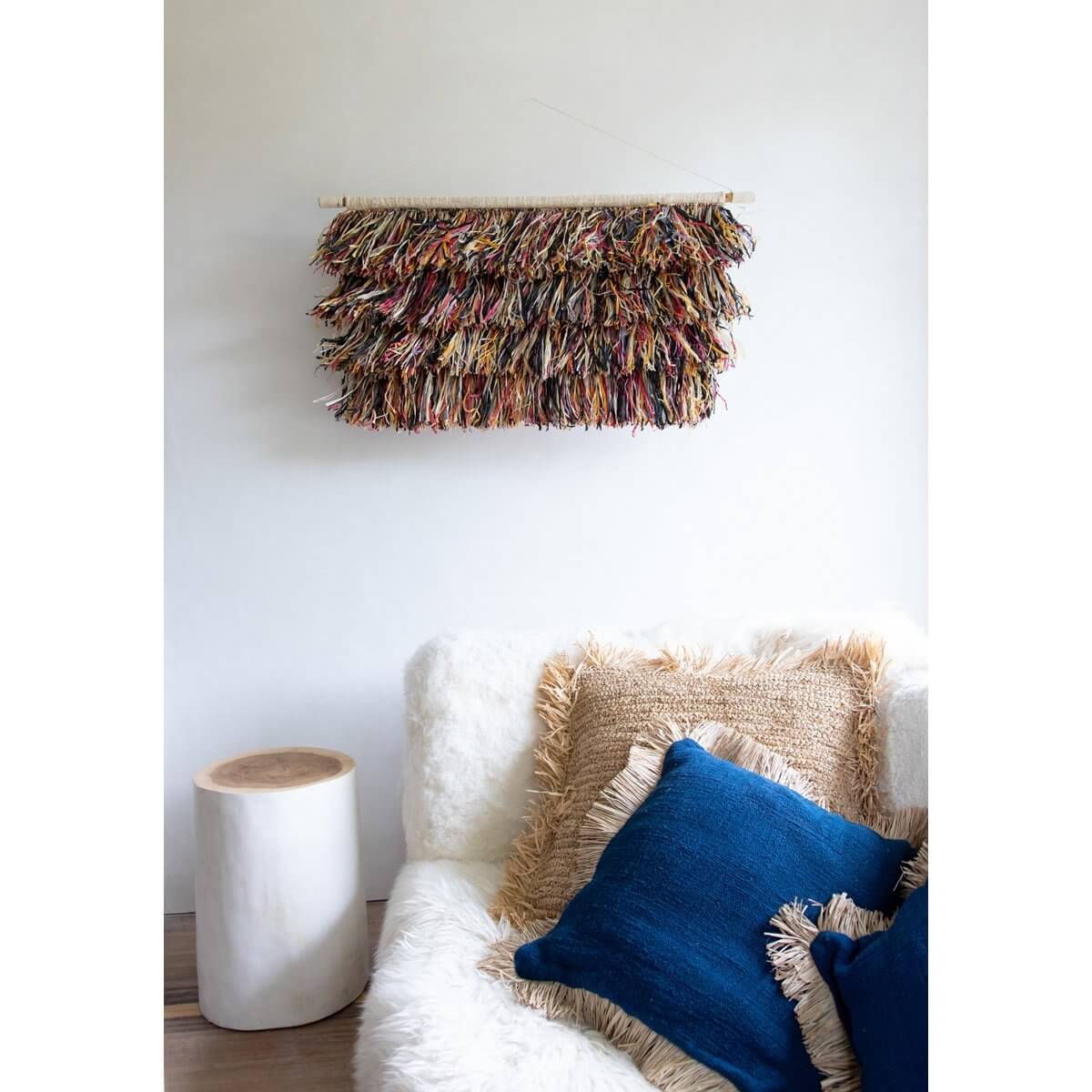 Boho chic interieur met blauwe en natuurlijke kussenhoes, raffia muurdecor en houten bijzettafel. Perfect voor tropische vibes.