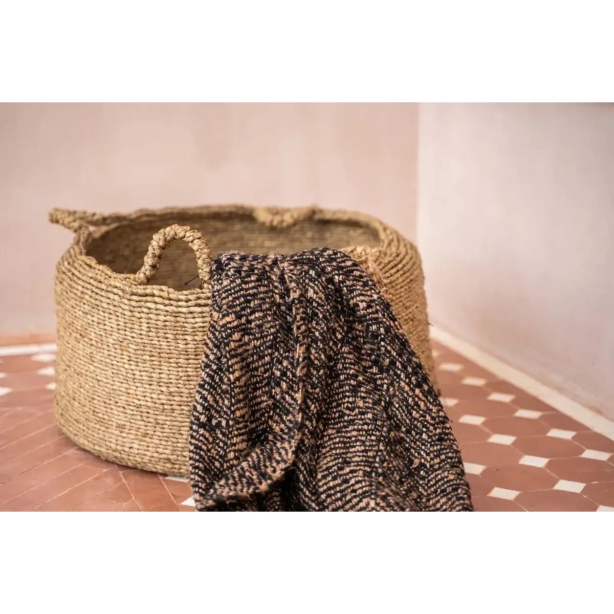 Handwoven sea grass basket with four handles, holding a textured blanket on a patterned tile floor. Perfect for stylish storage.
